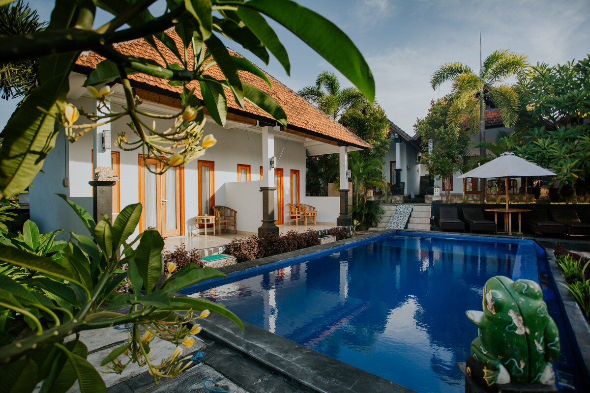 A serene outdoor area is depicted, featuring a clear swimming pool surrounded by lush greenery. Comfortable seating is arranged beside the pool, while a shaded table is positioned under a parasol. The bungalow displays a traditional roof and offers multiple entrances framed by sleek wooden doors.