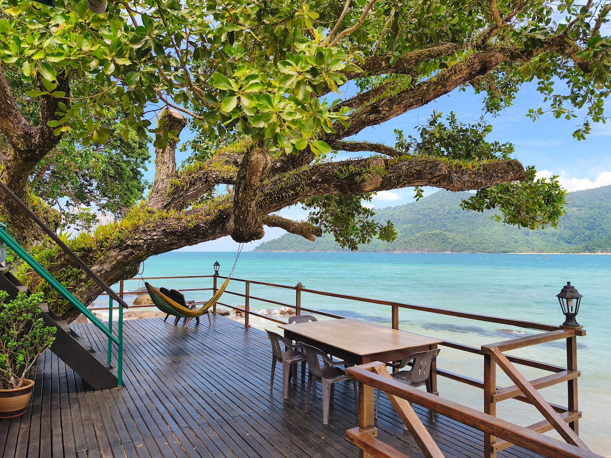 An outdoor terrace features a wooden deck with a large dining table and several chairs. A hammock is suspended from a large tree branch, offering a relaxing view of the turquoise sea and distant hills. A soft breeze is suggested by the gentle movement of the leaves.