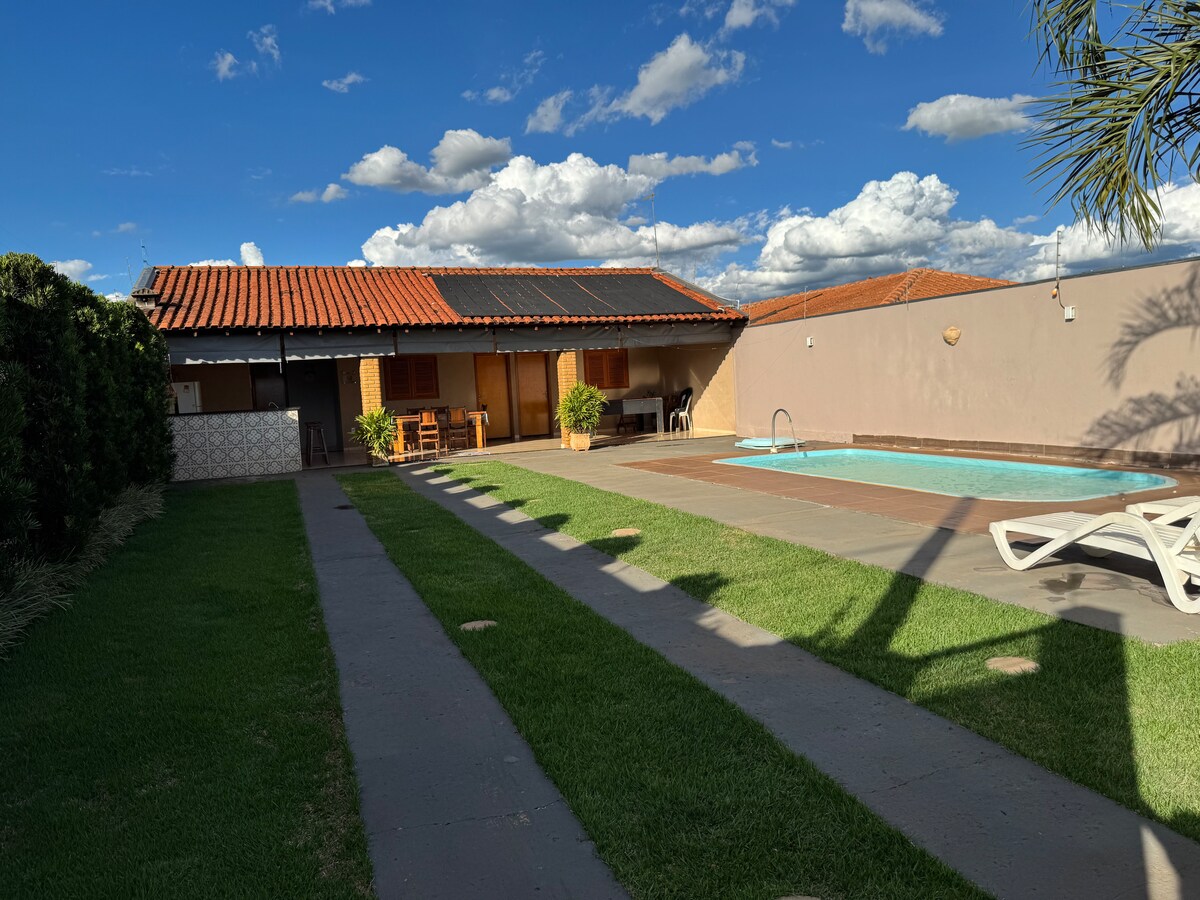 A single-story house features a red-tiled roof and wooden accents, surrounded by well-maintained grass. A private pool is visible alongside a patio area, with lounge chairs positioned for relaxation. Lush greenery borders the property, enhancing the serene outdoor environment.