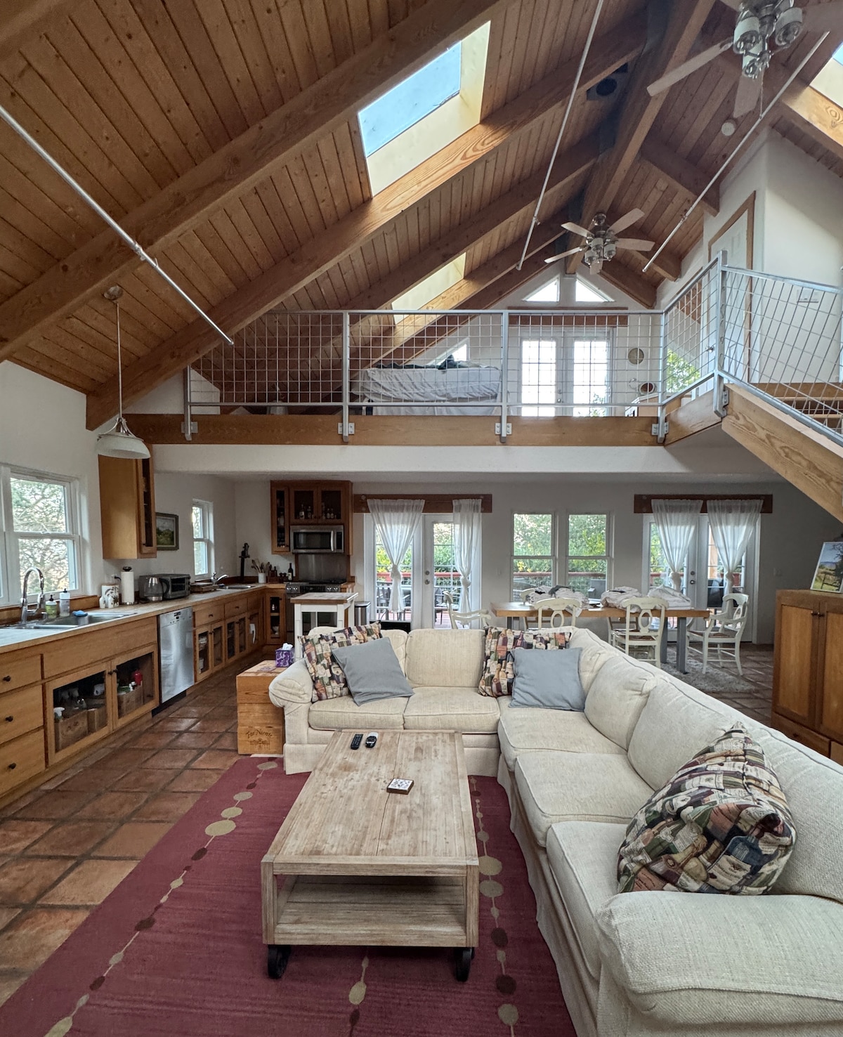 An open living area showcases a light-filled space featuring a comfortable sectional sofa, a wooden coffee table, and a well-equipped kitchen. The high ceiling with skylights adds to the airy feel, and large windows provide views of the outdoor surroundings.