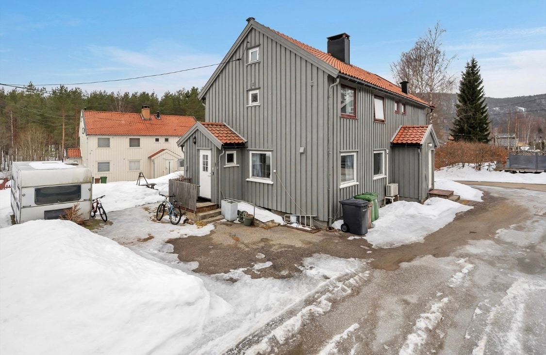 An exterior view of a wooden house with light gray siding and a red roof, situated next to snow-covered ground. Nearby, other buildings are visible, and a narrow road leads into the scene, with a mix of seasonal vegetation and snow at the edges.