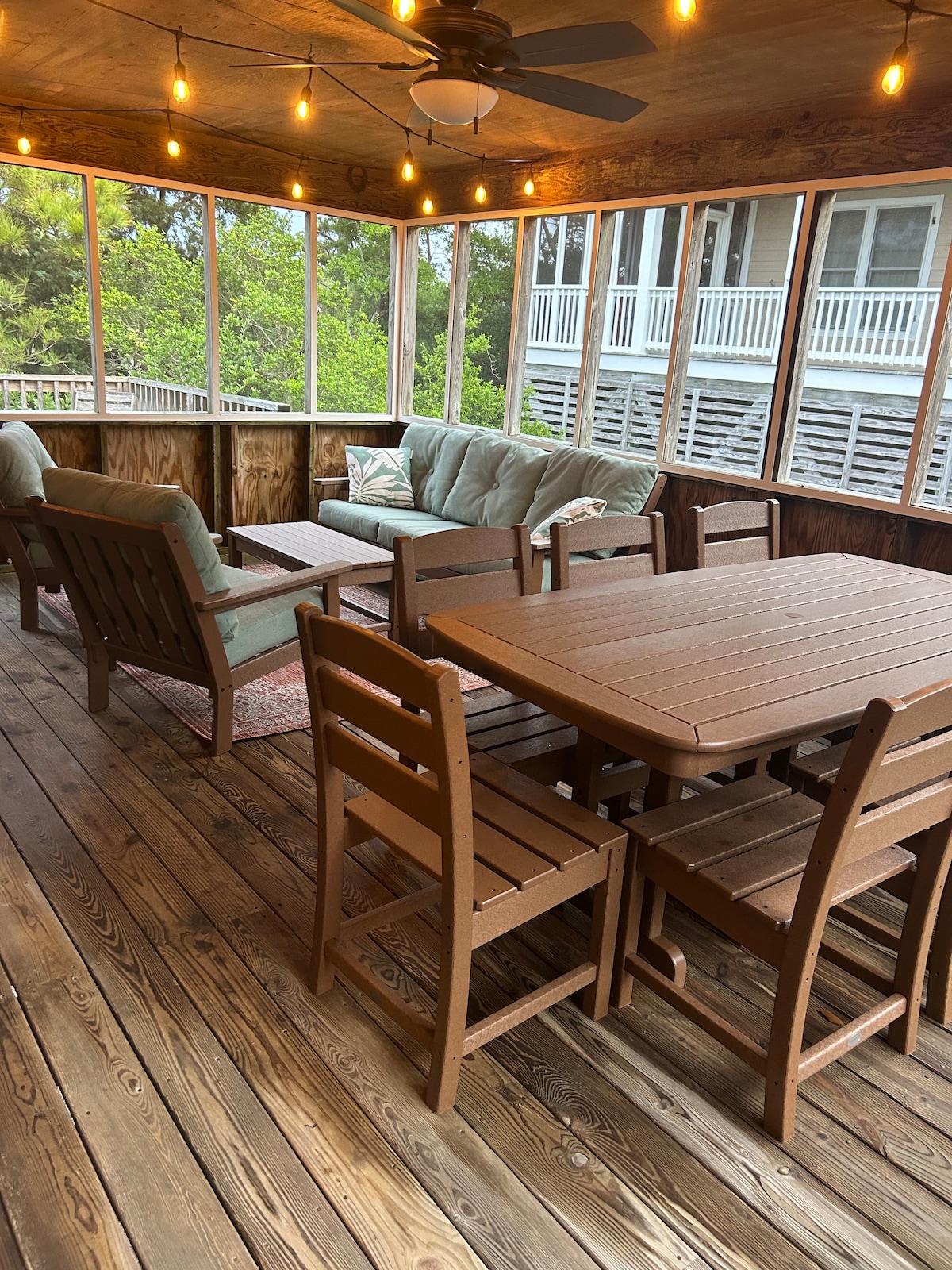 A furnished screened porch features a comfortable seating area with a plush sofa and armchairs, alongside a wooden dining table with seating for six. The space is illuminated by hanging lights and surrounded by natural wood paneling, creating a welcoming atmosphere.