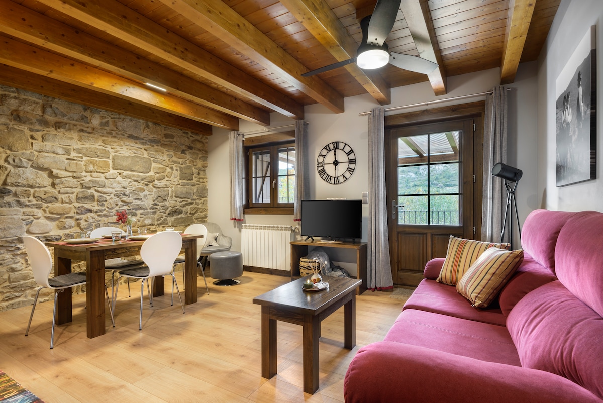 A living area features wooden beams and a stone accent wall, creating a cozy space. A large red sofa is positioned near a coffee table, with a dining table for four visible. Large windows provide natural light and a view of the outdoors.