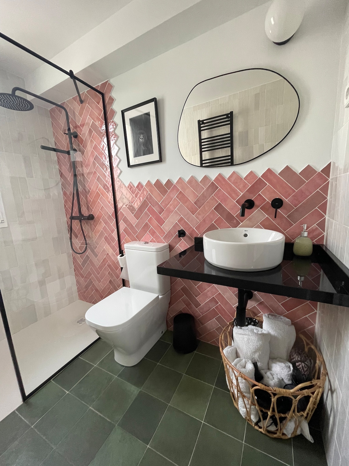 A modern bathroom features stylish pink and white tiled walls, complemented by a sleek black countertop with a circular sink. A rain shower is enclosed by a glass panel, while a framed artwork is displayed above. Soft towels are neatly arranged in a woven basket.