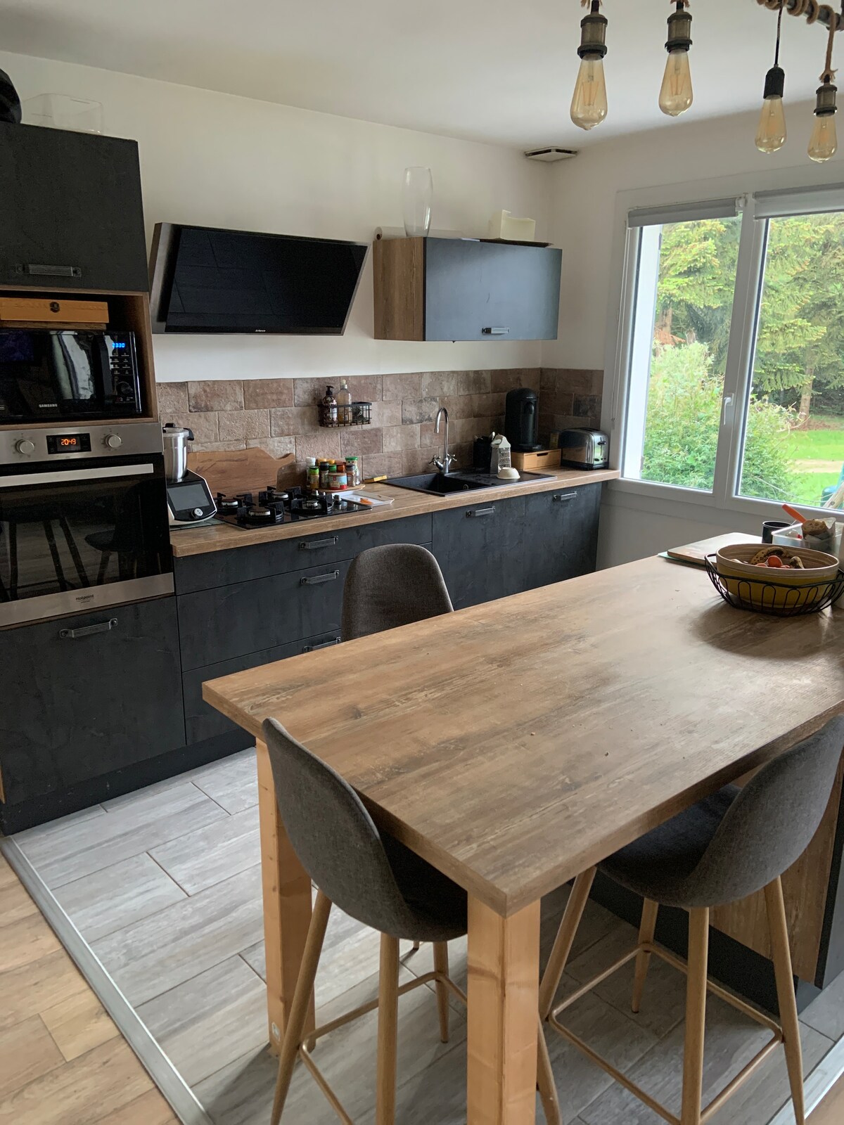 A modern kitchen features dark cabinetry and a light-colored wood dining table with three gray chairs. Kitchen appliances are integrated, and a large window provides natural light, overlooking a green outdoor area. Decorative items are arranged on the countertop, enhancing functionality.