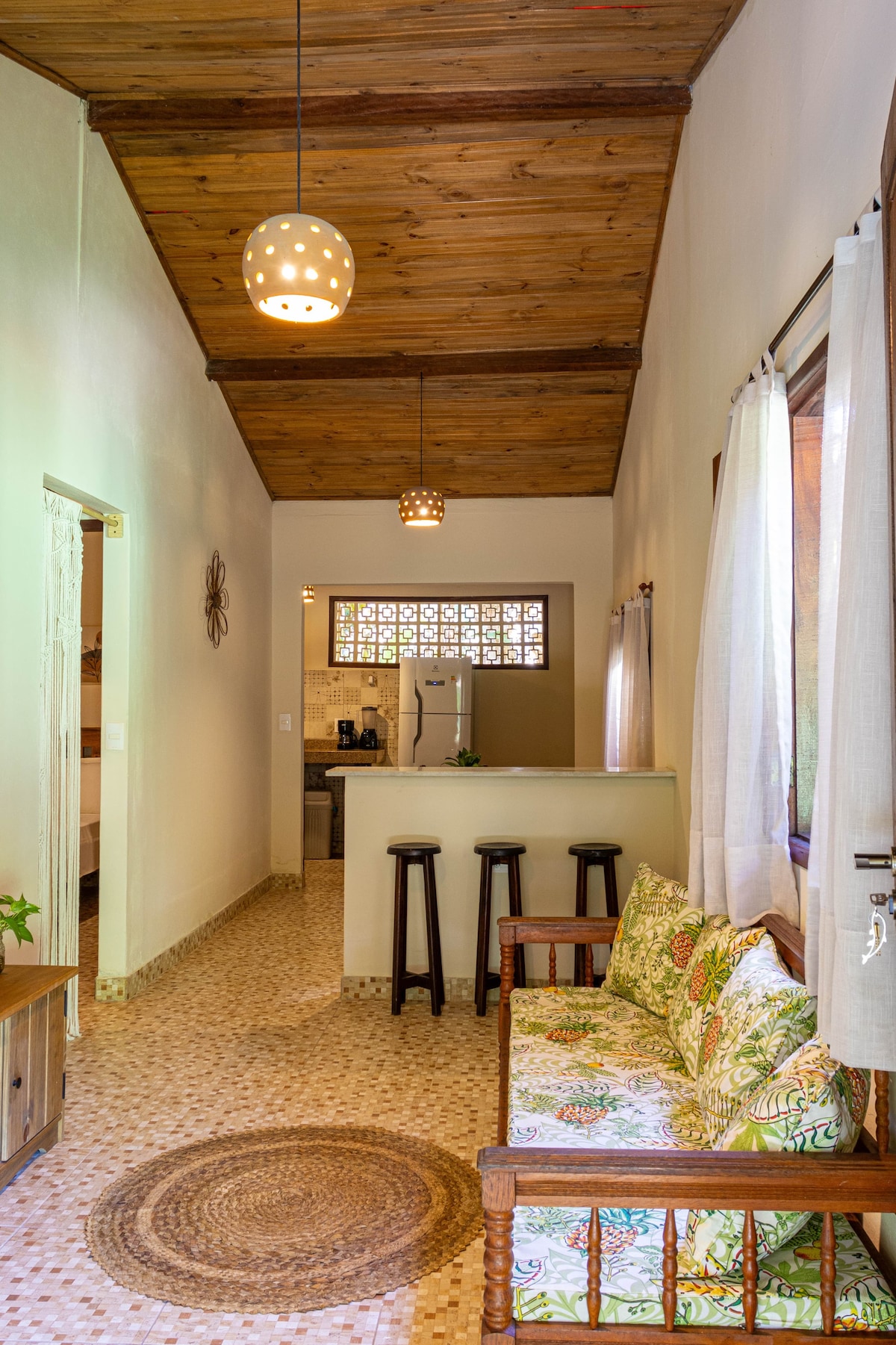 A cozy living area features a wooden bench adorned with floral cushions, offering a welcoming seating option. The space is illuminated by pendant lights, with a view toward the kitchen area where modern appliances are visible. Natural light fills the room through large windows.