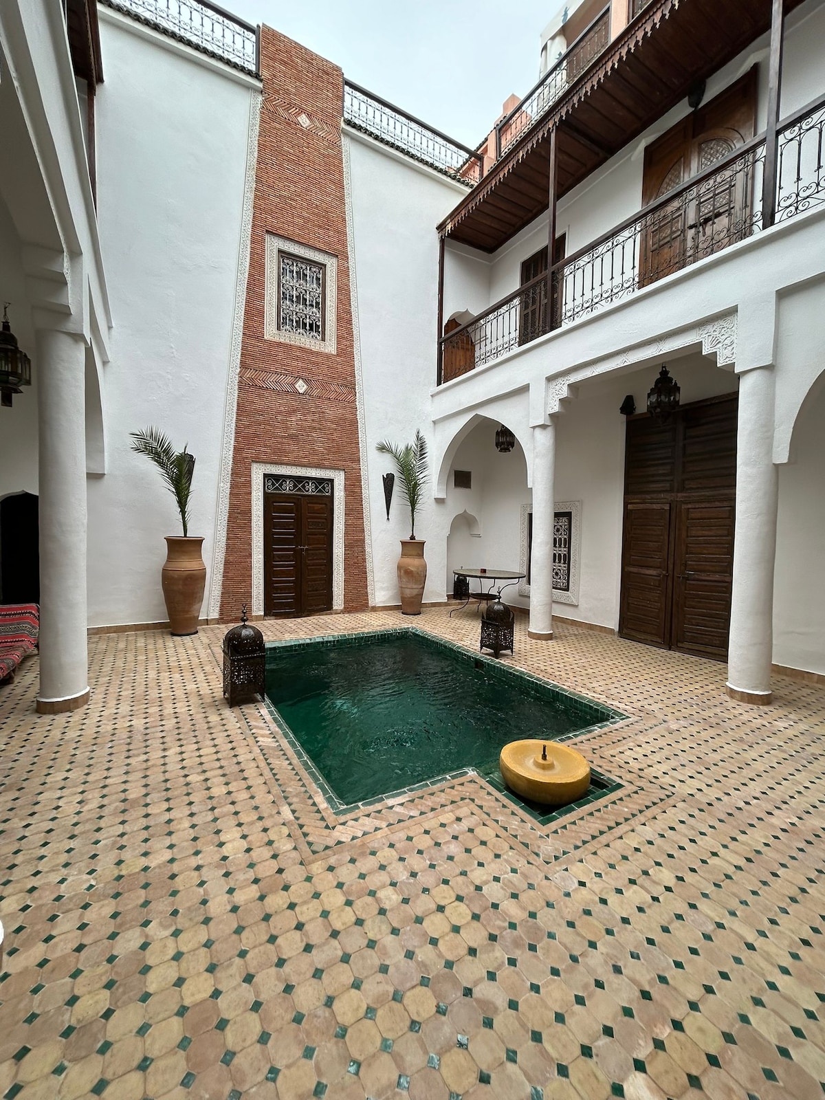 A central patio features a small pool with dark green water, surrounded by patterned tiles. Tall clay pots and palm plants add a touch of greenery, while wooden doors and decorative arches frame the space, creating a serene and inviting atmosphere.