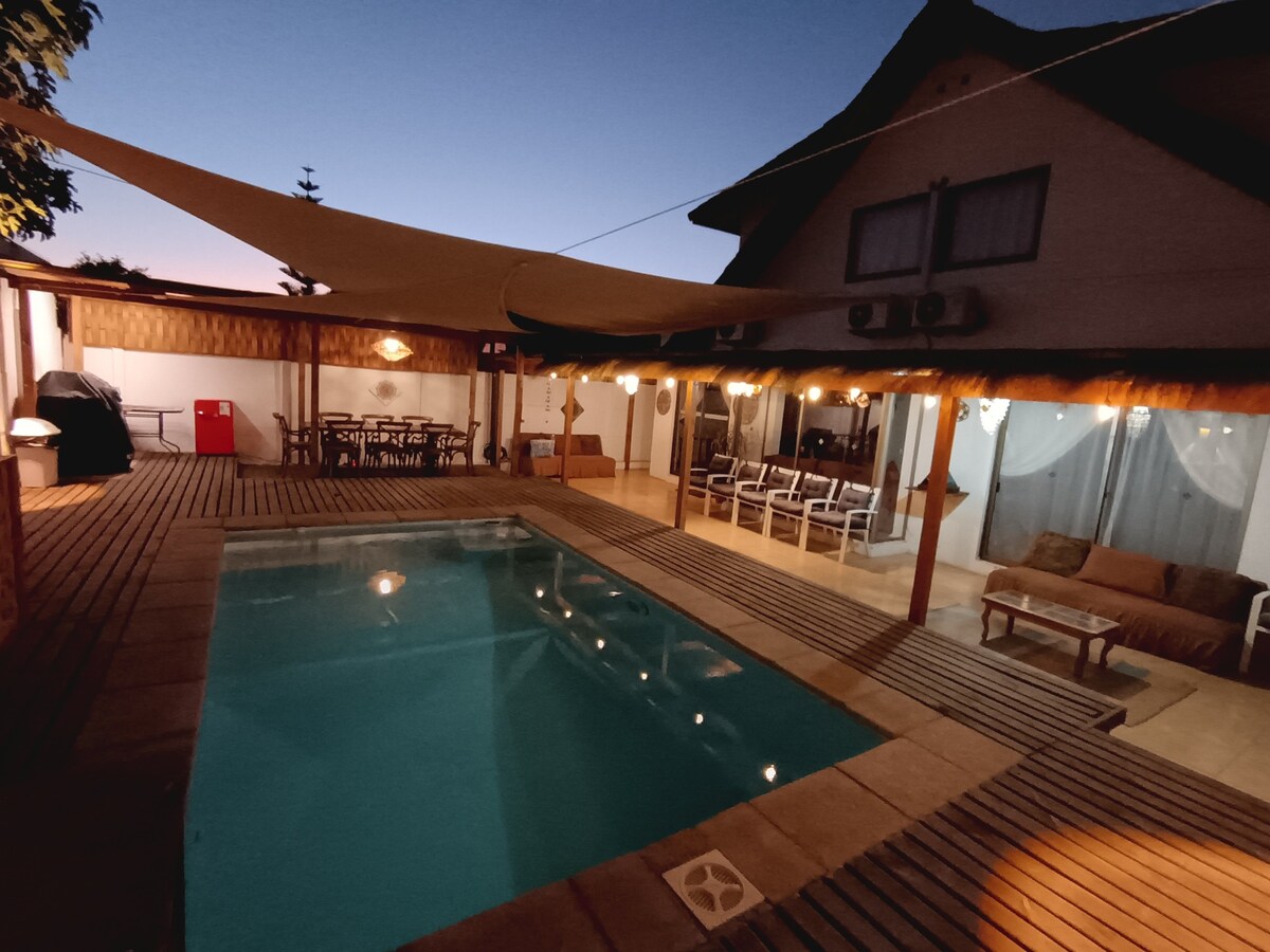 An outdoor patio area is highlighted by a private pool surrounded by wooden decking. Seating areas are present, including a shaded dining space with a large table. Soft lighting enhances the ambiance of the space, while the house is visible in the background.