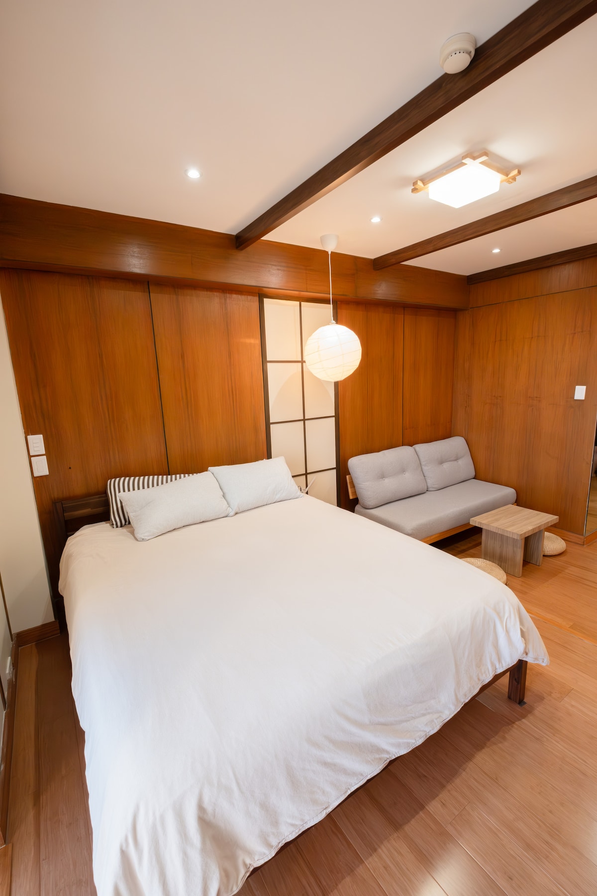 A cozy sleeping area features a spacious bed with a light-colored duvet and several pillows. A minimalistic gray daybed with a small wooden table is positioned beside it, and warm wooden paneling accentuates the walls, complemented by soft overhead lighting.