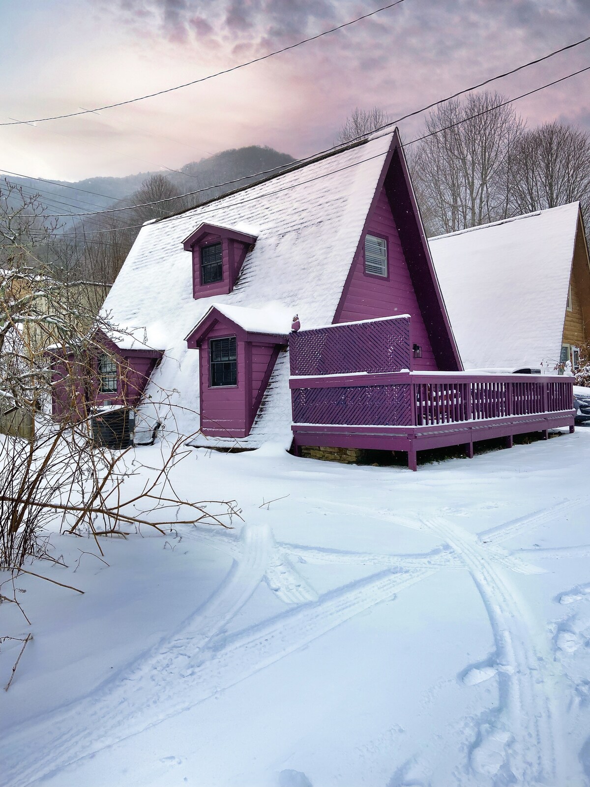 A vibrant purple A-frame chalet stands amidst a fresh blanket of snow, surrounded by leafless trees. A spacious deck can be seen at the front, featuring wooden railings. Snow-covered pathways lead towards the entrance, creating a serene winter scene.