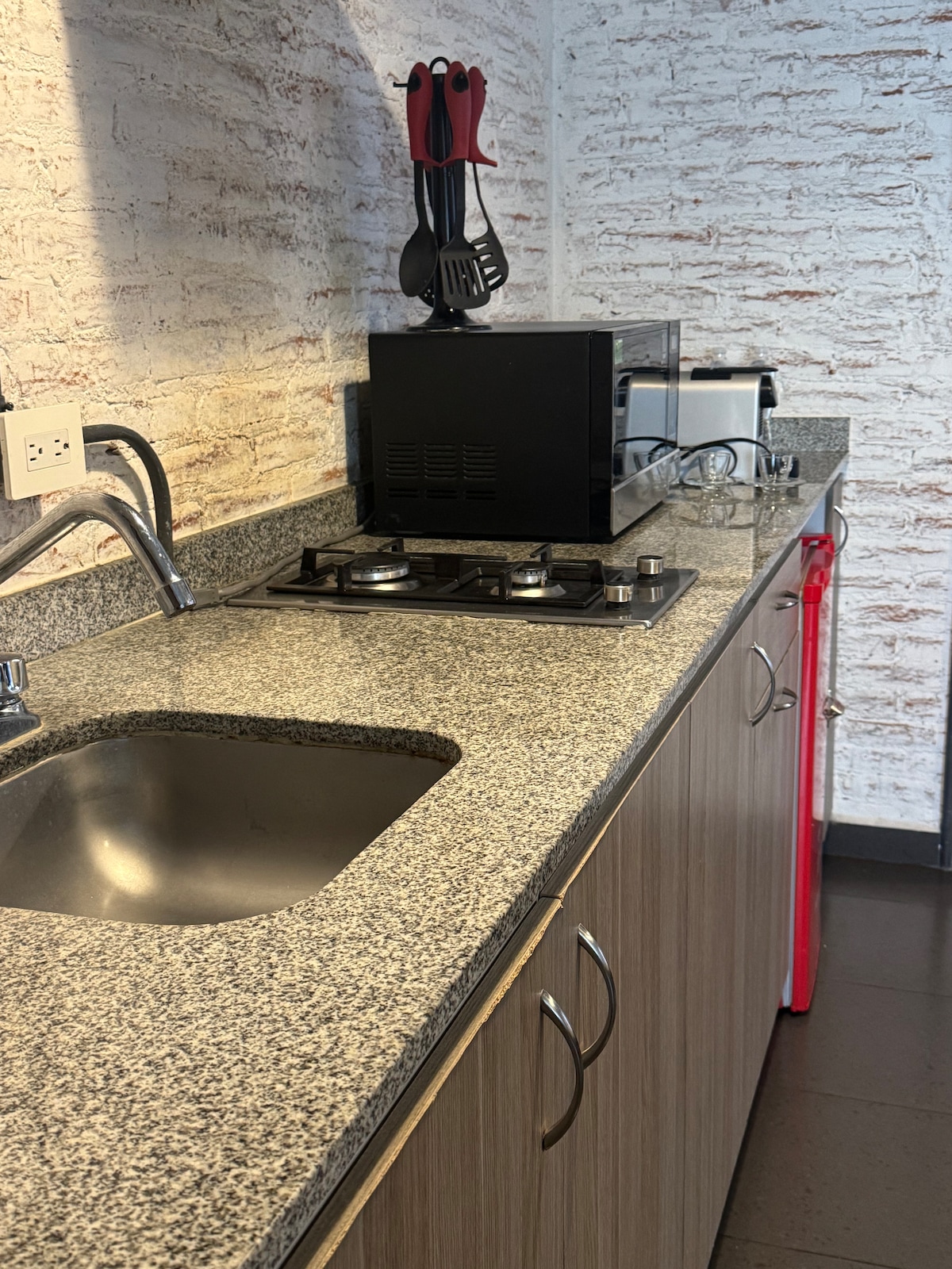 A compact kitchen space is equipped with a sleek granite countertop, featuring a stainless steel sink and a gas stove with two burners. A microwave and toaster are placed beside the black kitchen unit, while utensils are organized in a holder on the wall.