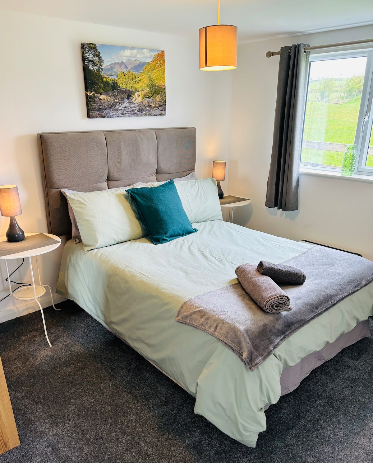 The bedroom is furnished with a spacious bed featuring a soft headboard. Two bedside tables are positioned alongside the bed, each with a lamp. A vibrant landscape artwork decorates the wall above the bed, and natural light enters through a window covered by matching grey curtains.