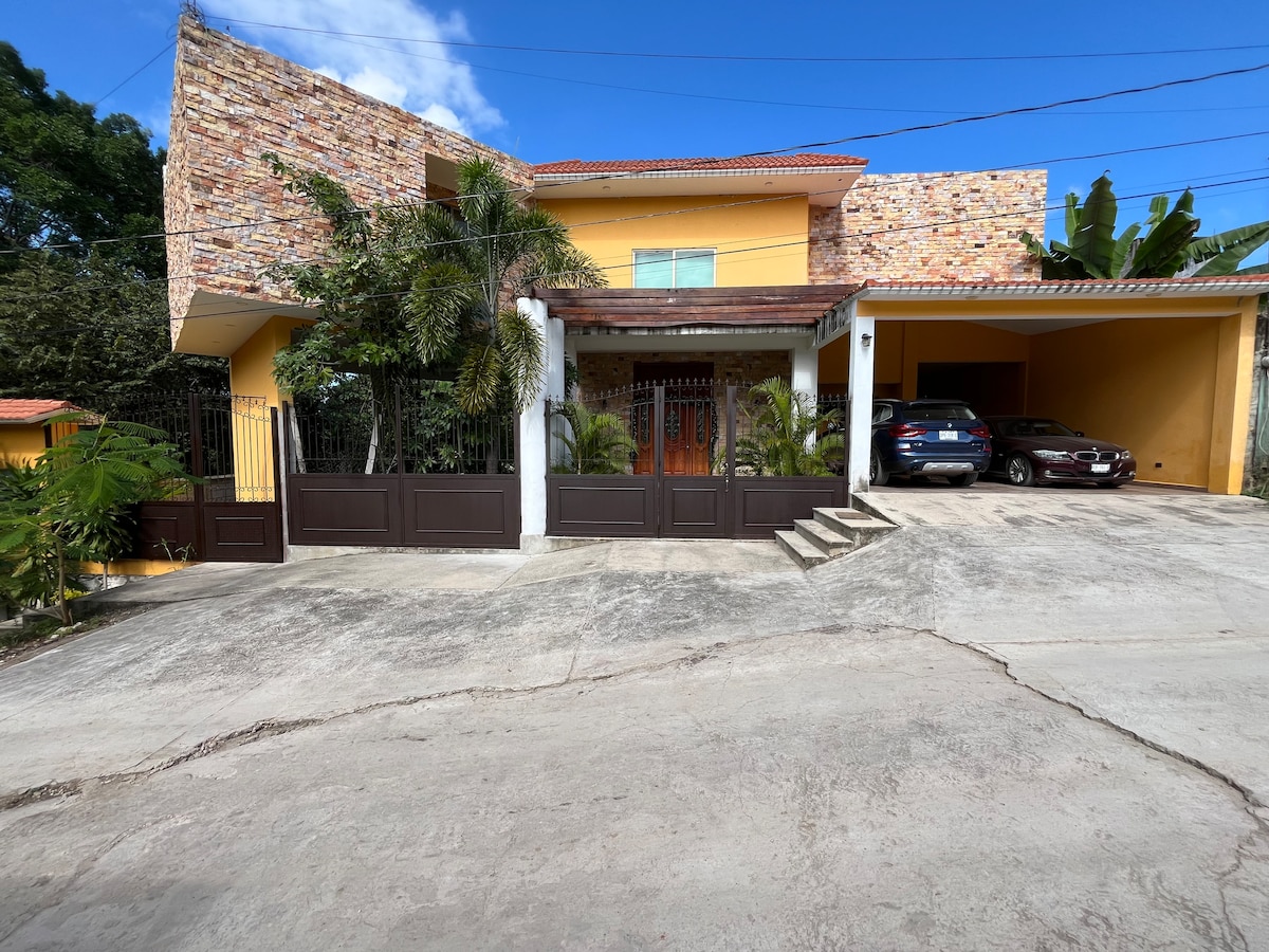 The exterior of a two-story building is presented, featuring a combination of stone and yellow walls. A landscaped entrance with palm trees and a gated driveway is visible. Two cars are parked in the driveway, and bright blue skies are seen above.