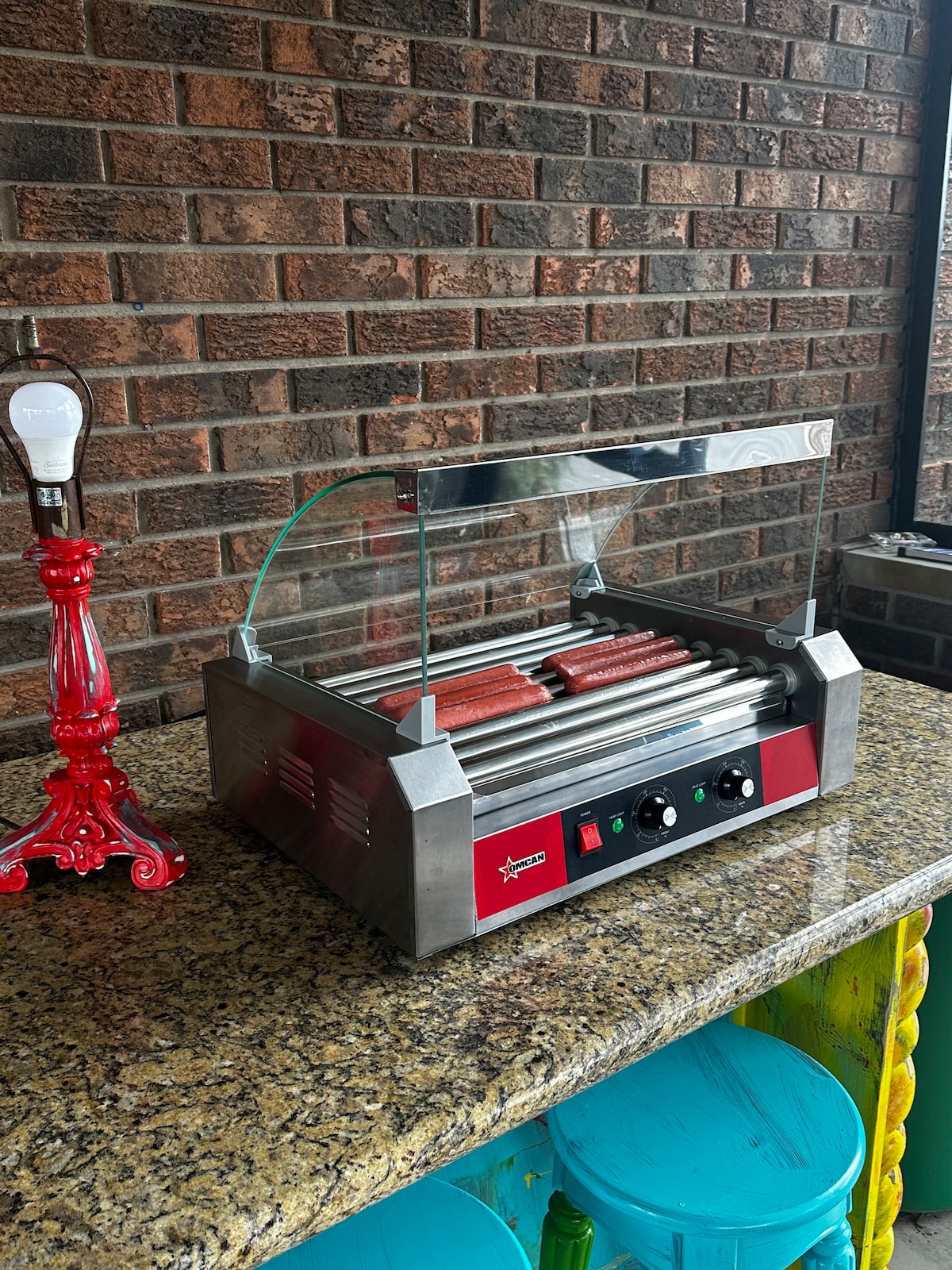 A hot dog roller grill is displayed on a granite countertop. The grill features multiple heated rollers and control knobs, with a glass shield protecting the food. A decorative red tap is visible beside the grill, adding to the outdoor cooking area.