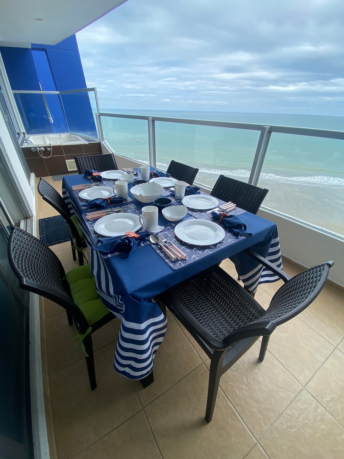 A dining table is set on a balcony overlooking the ocean. The table is covered with a blue and white striped tablecloth and arranged with white plates, mugs, and utensils. Surrounding chairs provide comfortable seating, with the ocean visible through glass railings.