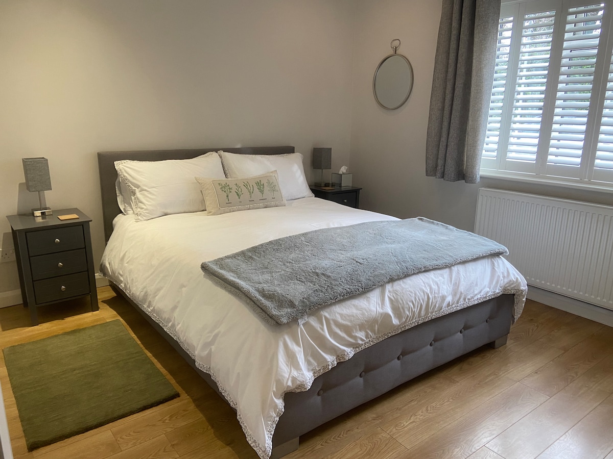 A spacious bedroom features a large bed with fresh white bedding and a light blue throw. Dark wood nightstands are positioned on either side, each with a lamp. Natural light enters through the shuttered window, and a round mirror is mounted on the wall.