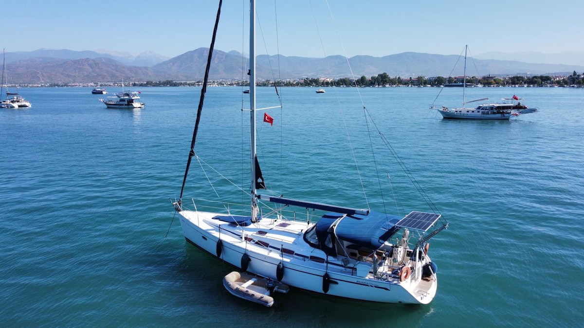 A sailboat is anchored in calm, turquoise waters, surrounded by a scenic coastal backdrop. The vessel features a covered seating area and solar panels, with paddle boards positioned on its side. Nearby boats can be seen in the distance under a clear blue sky.