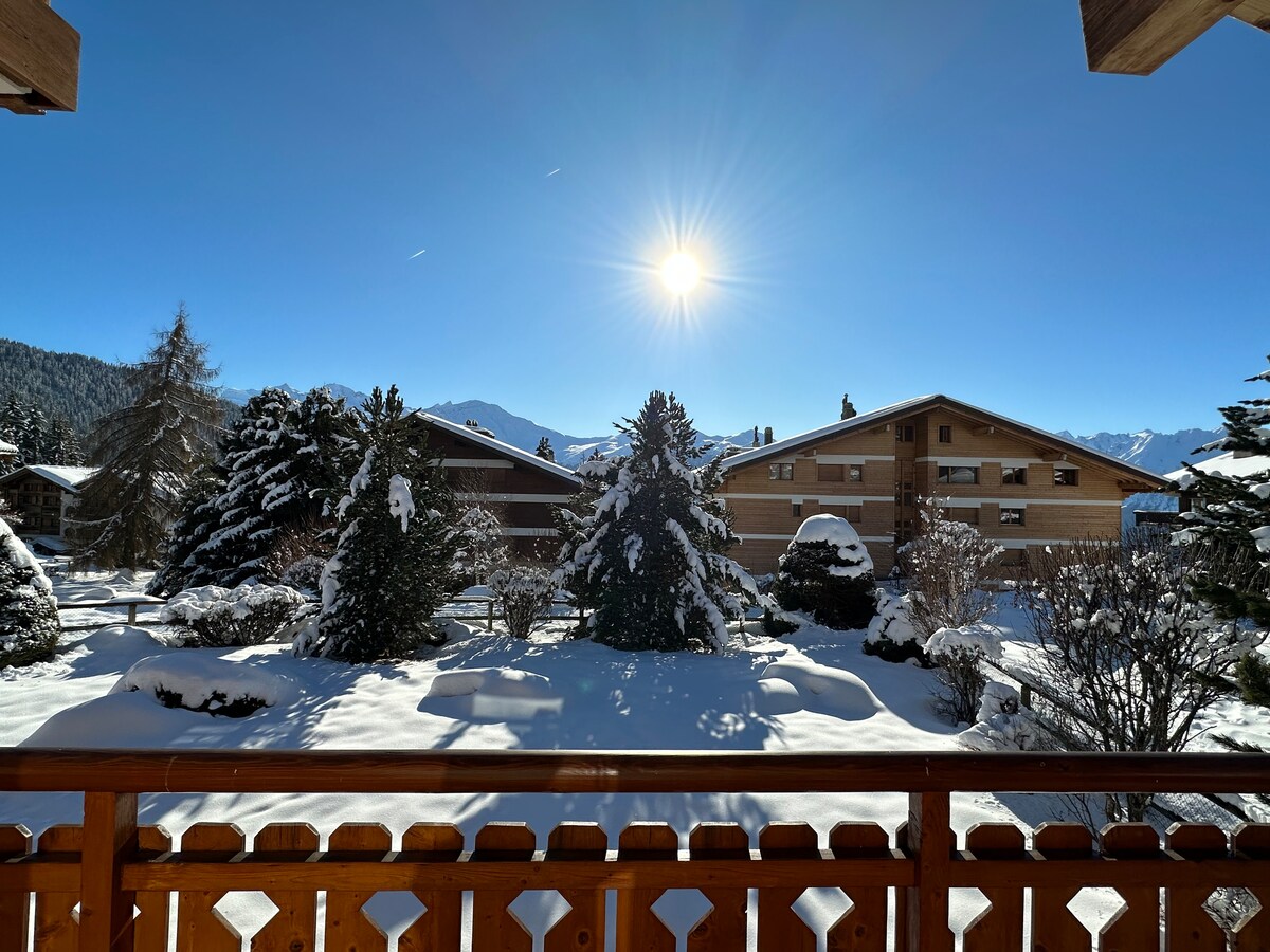 A winter view from a balcony captures a serene landscape blanketed in fresh snow. Sunlight shines brightly in the clear blue sky, casting shadows on nearby evergreen trees and snow-covered shrubs. Traditional chalets are nestled among the snowy surroundings, enhancing the tranquil scene.