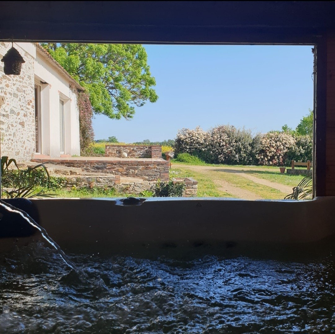 A view of an outdoor landscape is framed through an open window, showcasing a lush garden with flowering shrubs in full bloom. A calming spa with bubbling water is visible in the foreground, inviting relaxation and connection with nature.