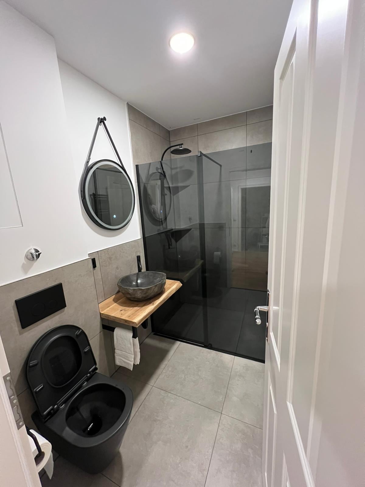 A modern bathroom is presented with sleek gray tiles and minimalist design elements. A frameless glass walk-in shower is visible, alongside a vessel sink atop a wooden countertop. Two round mirrors are mounted on the wall, and a compact black toilet is positioned within the space.