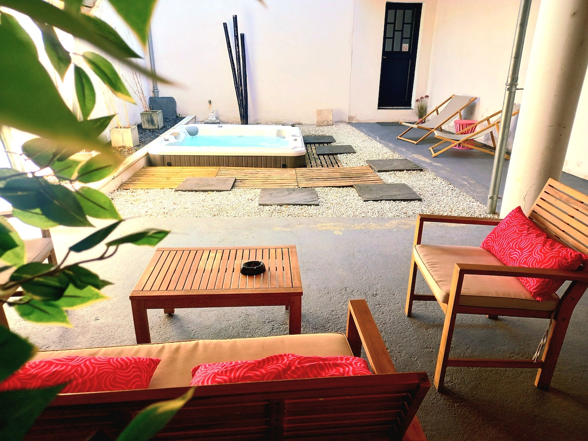 A private patio area is showcased, featuring a Sundance Spa surrounded by wooden decking and laid stones. Comfortable wooden furniture with red-accented cushions is present. A door in the background leads to another part of the property, adding a sense of seclusion.