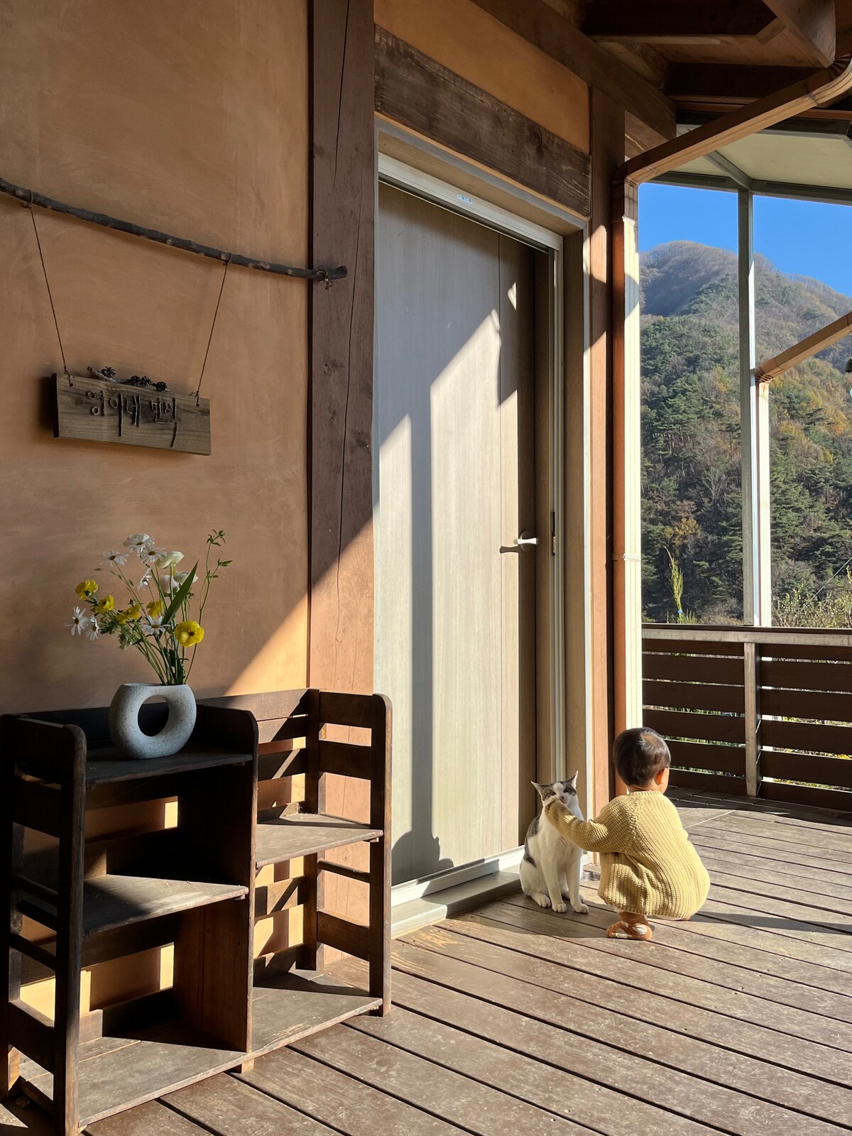 A cozy entrance features a wooden door leading to a tranquil balcony. A child kneels beside a friendly cat, showcasing a moment of connection. A small flower arrangement adorns a shelf nearby, adding a touch of nature. The backdrop reveals a scenic mountain view.