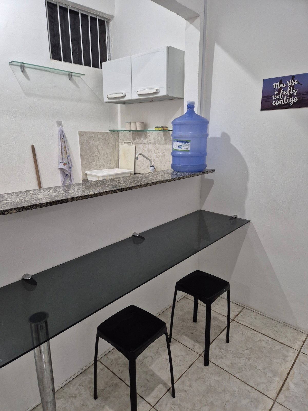 A clean kitchenette area is presented, featuring a granite countertop with a sink. Two black chairs are positioned at a glass-top table. A water dispenser is visible on the counter, with light illuminating the space through a small window.