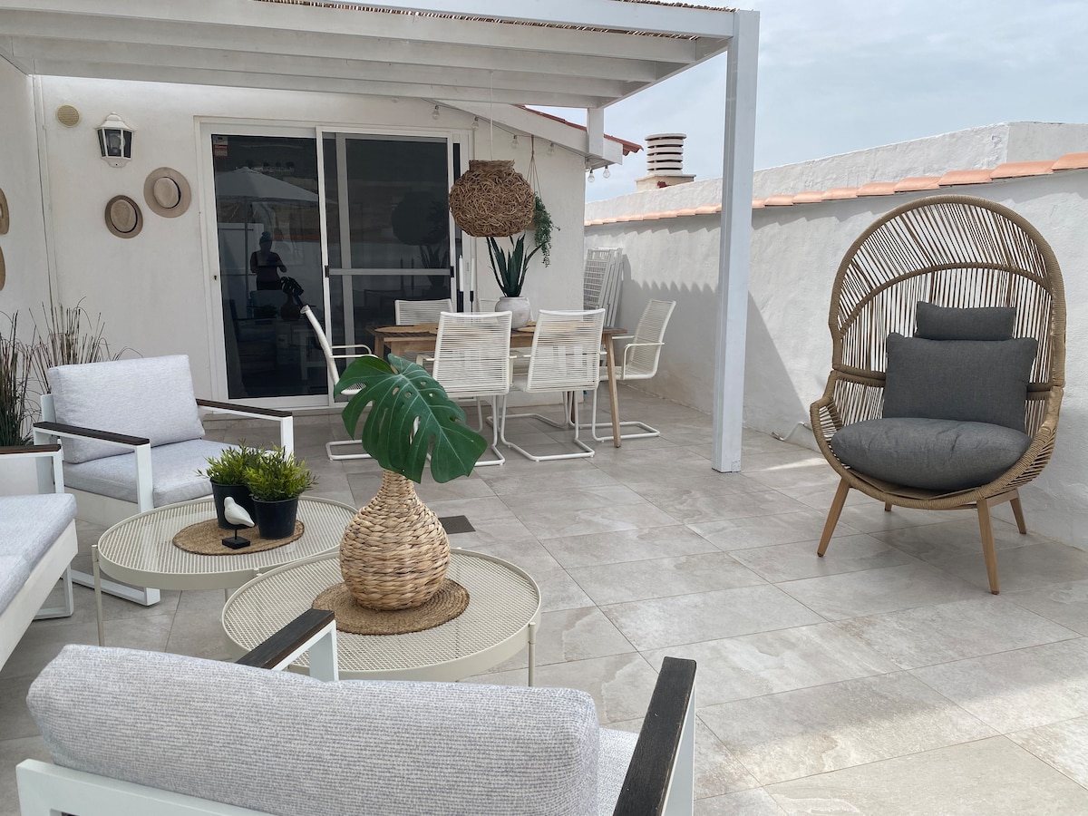 An outdoor area features a comfortable seating arrangement, including a circular chair and white lounge chairs surrounding a table. A wooden dining table is visible in the background, with potted plants adding a touch of greenery. The space is covered by a white pergola, providing shade.