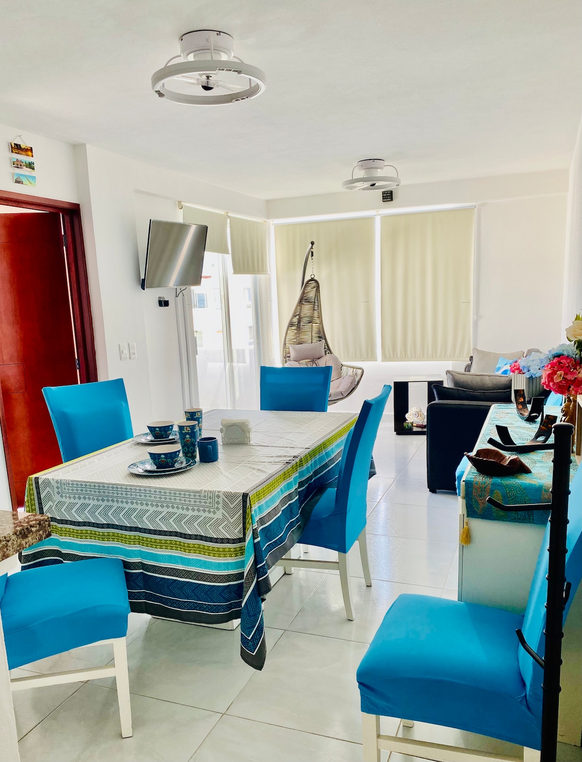 A dining area is showcased with a light-colored table set for four, featuring blue chairs and a decorative tablecloth. A hanging chair is visible in the background, with natural light filtering through large windows adorned with light blinds. Wall-mounted television enhances the modern ambiance.
