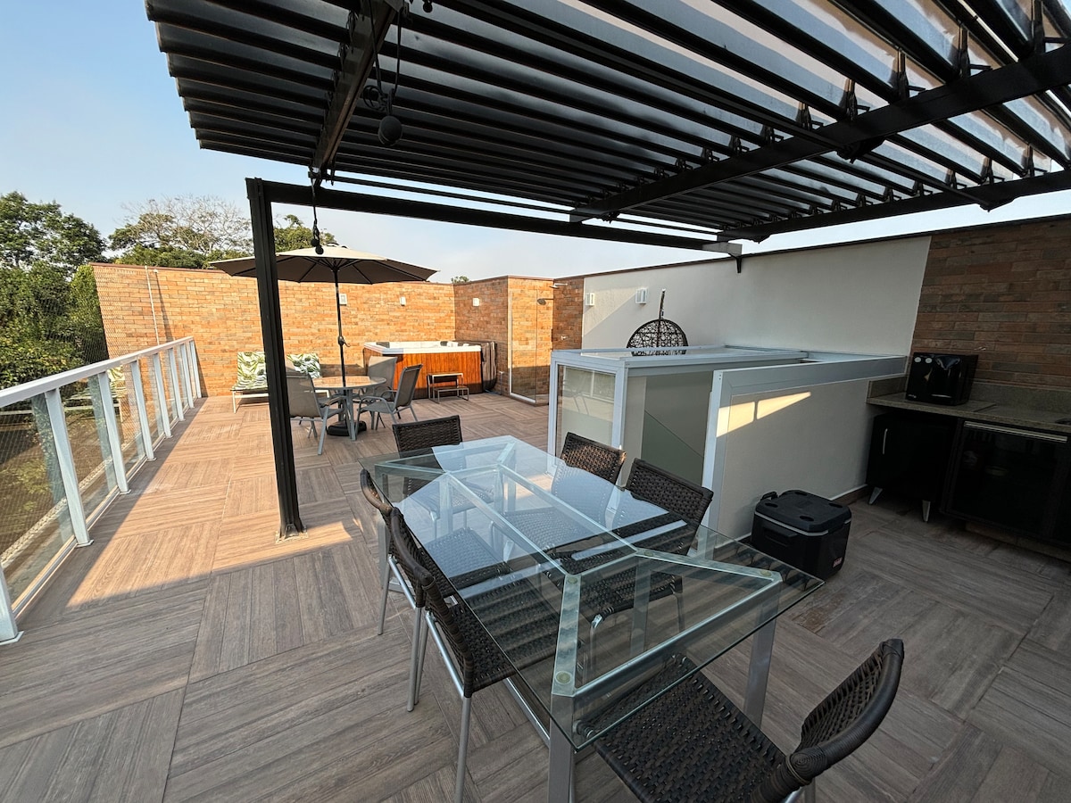 An outdoor terrace features a glass dining table surrounded by wicker chairs. A shaded area includes an outdoor kitchen and seating arrangement. The space is bordered by brick walls, with a clear view of the sky. A barbecue grill is situated near the dining area.