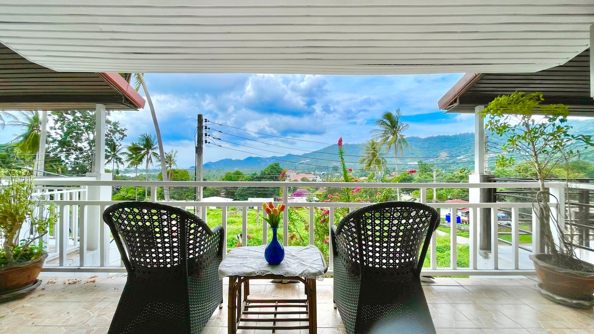 An outdoor balcony features two black wicker chairs positioned around a small wooden table. A vase of flowers sits atop the table, with a scenic view of mountains and palm trees in the background. Soft natural light illuminates the space.