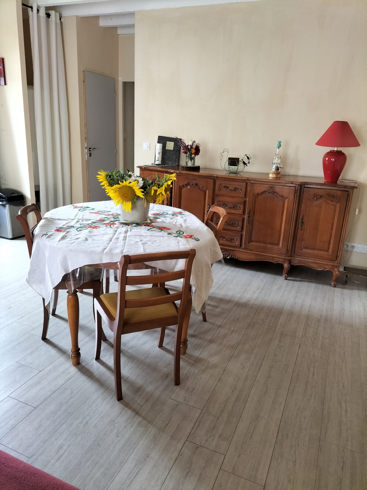 A dining area features a round table covered with a floral tablecloth, complemented by four wooden chairs. A vase filled with sunflowers adds a touch of color. A vintage wooden cabinet stands against the wall, showcasing decorative items and a small red lamp.