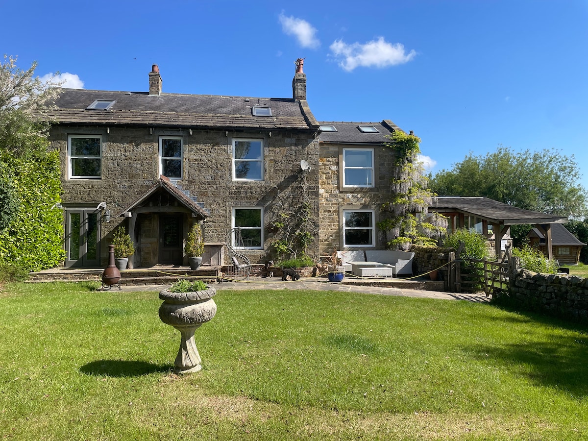 A two-story farmhouse is set against a bright blue sky, surrounded by lush green gardens. The stone facade is complemented by large windows, providing natural light. A spacious patio area features outdoor seating, while a decorative stone fountain adds an inviting touch to the landscape.