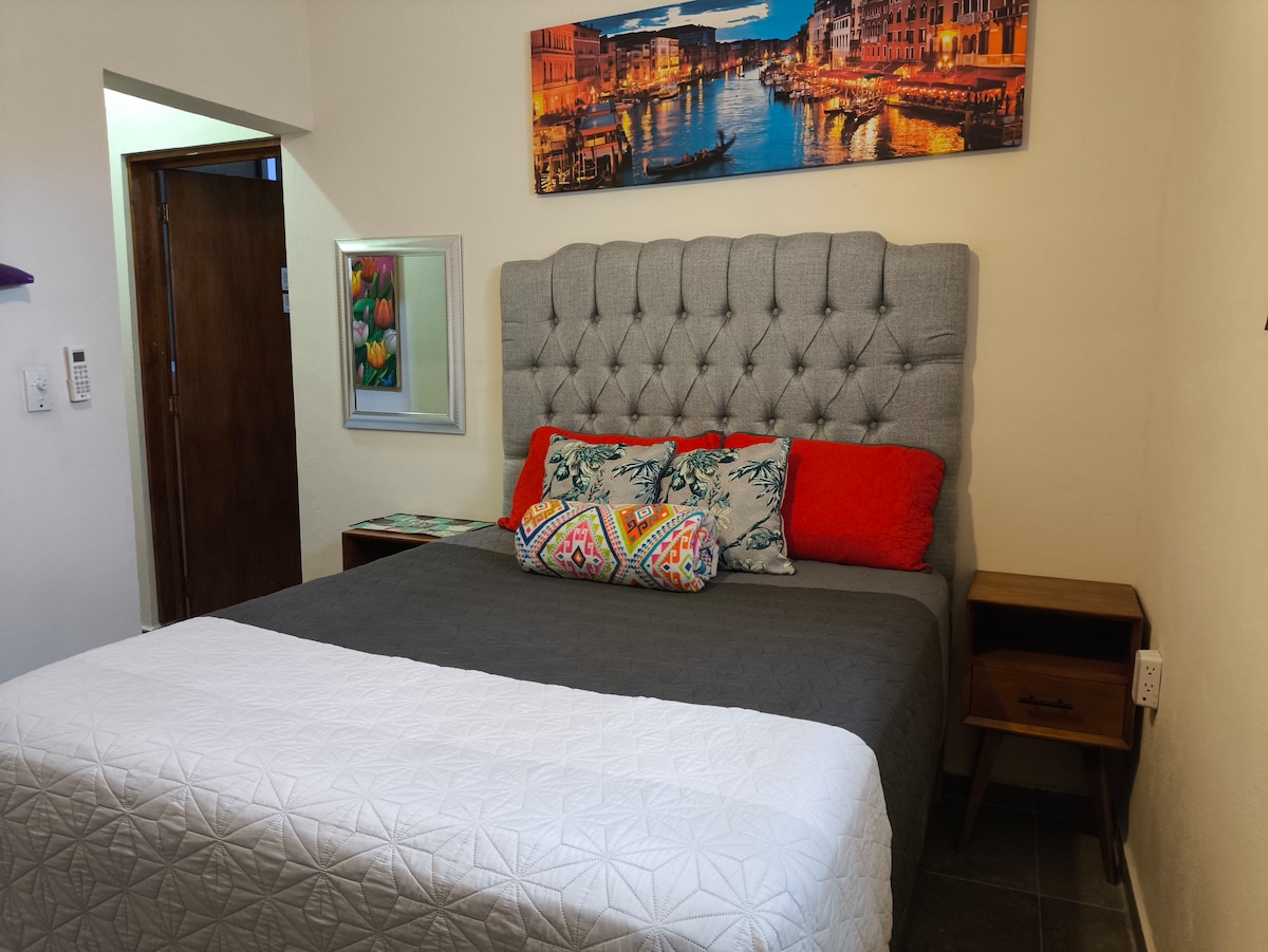 The spacious bed is dressed in neutral tones, featuring a tufted gray headboard and adorned with colorful decorative pillows. A small nightstand is positioned on one side, along with a framed mirror and wall art depicting a serene cityscape, enhancing the cozy atmosphere.