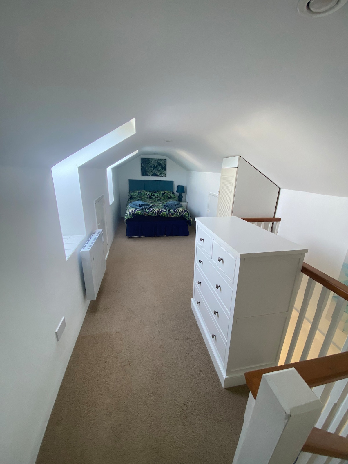 A well-lit upper landing is observed, featuring soft beige carpeting and minimalistic white walls. A spacious bed with a patterned duvet is situated in the background, complemented by a bedside lamp and a piece of wall art. A tall white dresser stands prominently in the foreground.