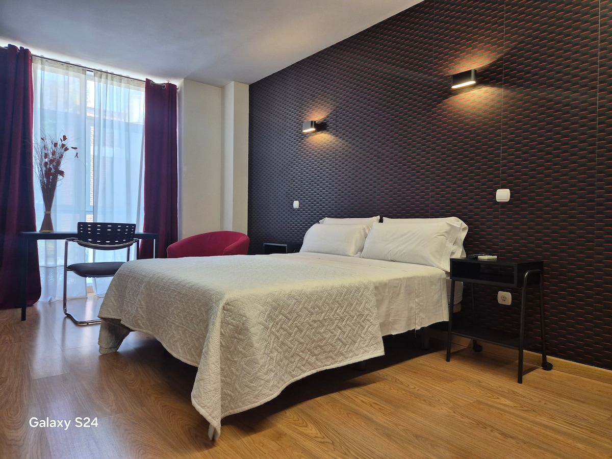 A modern studio space features a spacious bed with a white quilt and decorative pillows. A large window, dressed with sheer curtains, allows natural light to fill the room. A table and chair provide a functional workspace, and wall-mounted lights add illumination.