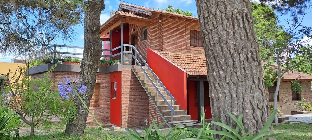 The exterior view reveals a two-story brick building featuring a vibrant red entryway and a staircase leading to the first floor. Surrounding greenery and tall trees create a serene environment, while a sunny sky enhances the inviting atmosphere of the location.