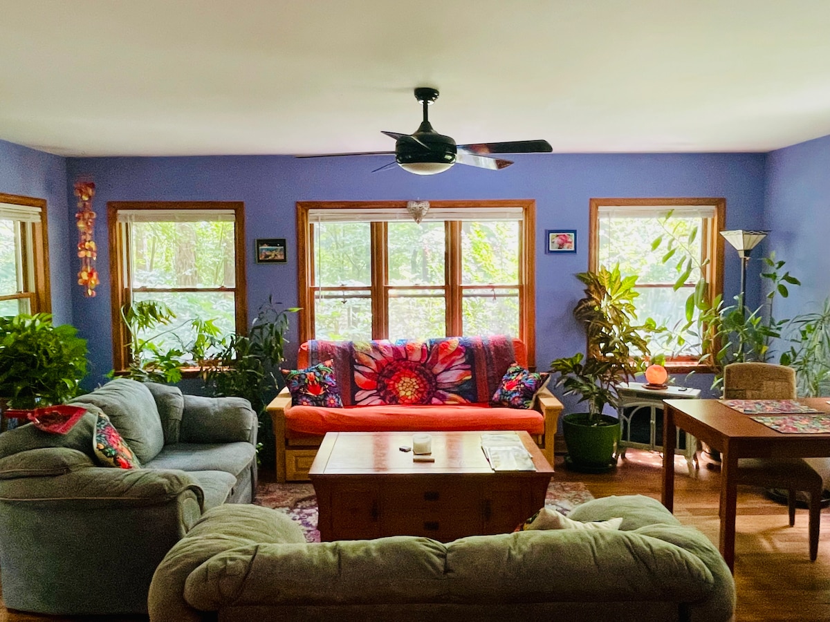 A spacious living area is surrounded by large windows, allowing natural light to illuminate the room. Soft furnishings, including a comfortable couch and loveseat, are arranged around a wooden coffee table. Lush green plants add a touch of nature, enhancing the serene atmosphere.