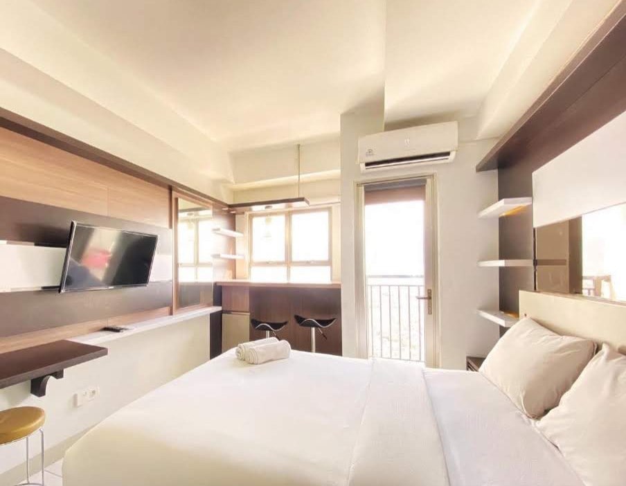 A comfortable room is shown featuring a spacious bed with soothing white linens. Natural light fills the space through large windows. Modern furnishings include a flat-screen TV mounted on the wall and stylish bar stools at a small table, enhancing functionality.