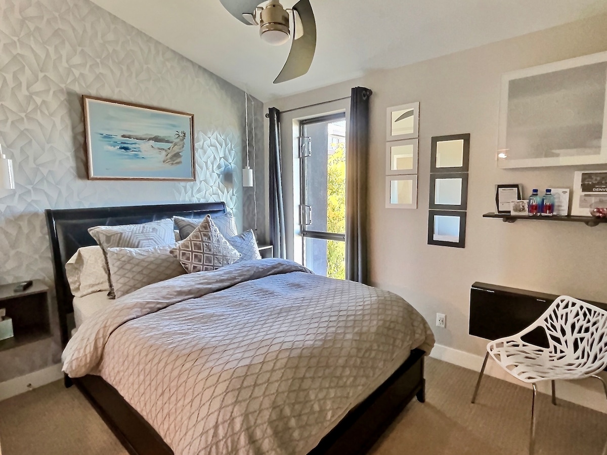 A private bedroom features a spacious bed with softly patterned bedding against a textured wall. Natural light enters through a window adorned with light curtains. A modern chair and wall-mounted shelves with bottled water are also visible, contributing to the room's functionality.