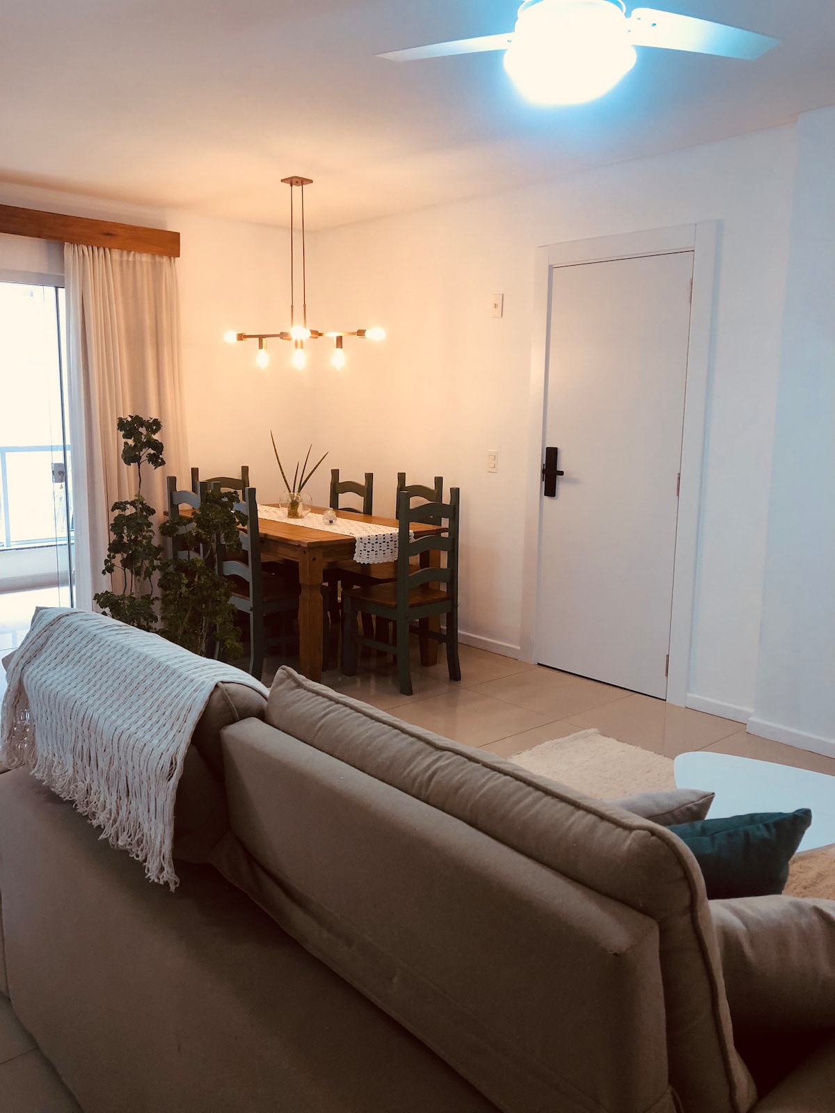 A dining area is visible featuring a wooden table surrounded by several chairs. A modern light fixture hangs above, illuminating the space. A cozy seating arrangement with neutral-toned cushions is positioned in the foreground, with a plant and window in the background.