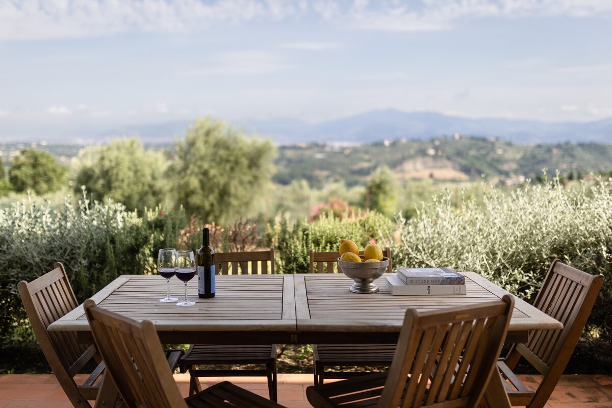 An outdoor dining area features a wooden table set with a fruit bowl and two glasses of wine. Surrounding olive trees create a natural backdrop, with distant hills visible under a clear sky. The setting invites relaxation and enjoying the scenic views.