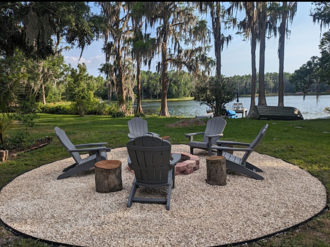A fire pit area is surrounded by four gray Adirondack chairs and natural wood stump seating, placed on a gravel surface. Lush greenery and tall trees frame the serene lake view in the background, creating a tranquil outdoor gathering space.