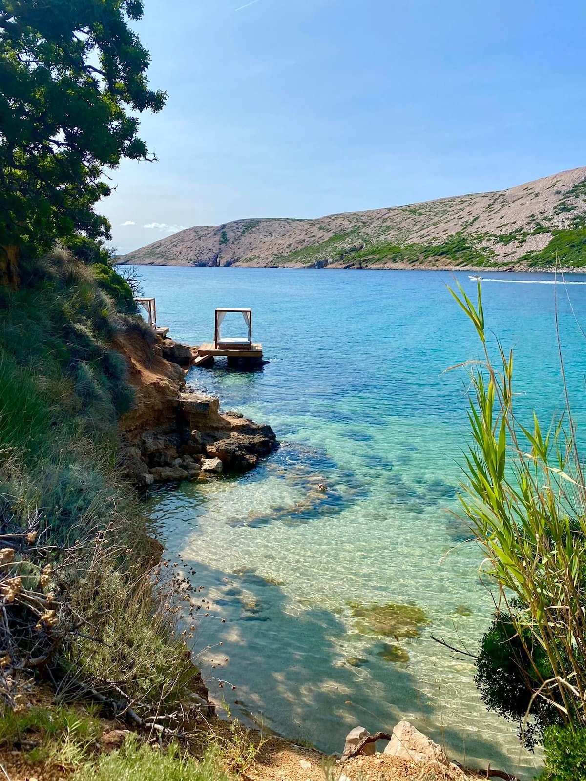 A serene coastal view showcases clear turquoise waters gently lapping against the rocky shore. Natural greenery frames the scene, with a wooden platform extending into the water. Surrounding hills provide a backdrop, emphasizing the inviting nature of the beach area.