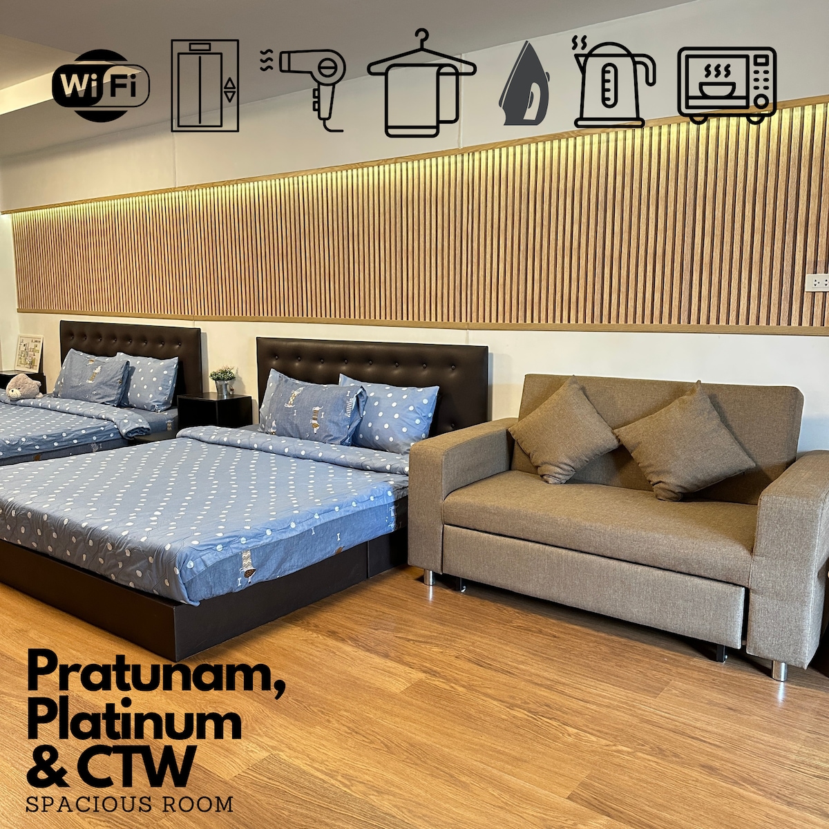 The room features two spacious king-size beds adorned with blue patterned bedding. A comfortable sofa is situated nearby, with wooden paneling on the wall enhancing the interior. Essential amenities are displayed above, including a WiFi symbol and various appliances.
