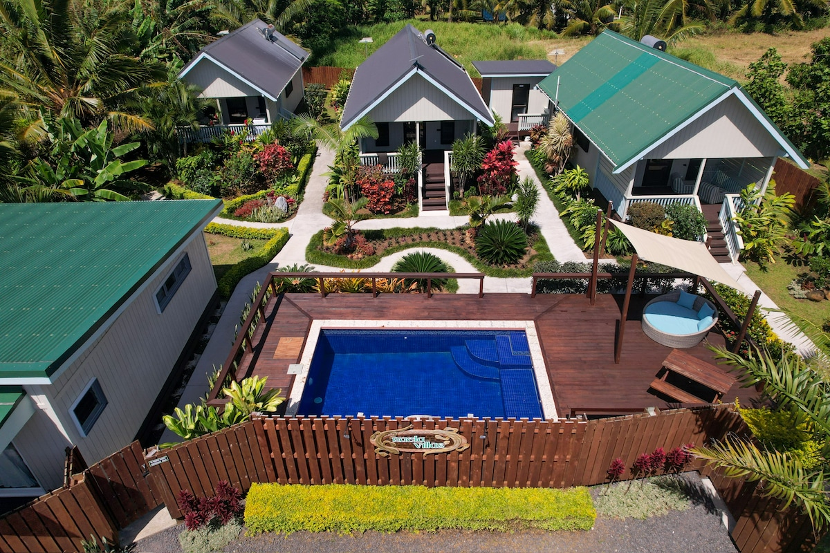 An aerial view showcases a landscaped courtyard featuring a private pool framed by wooden decking. Surrounding the pool, vibrant tropical plants and decorative elements enhance the serene environment. Three villas, each with distinctive roofs, are positioned harmoniously within the well-maintained garden.
