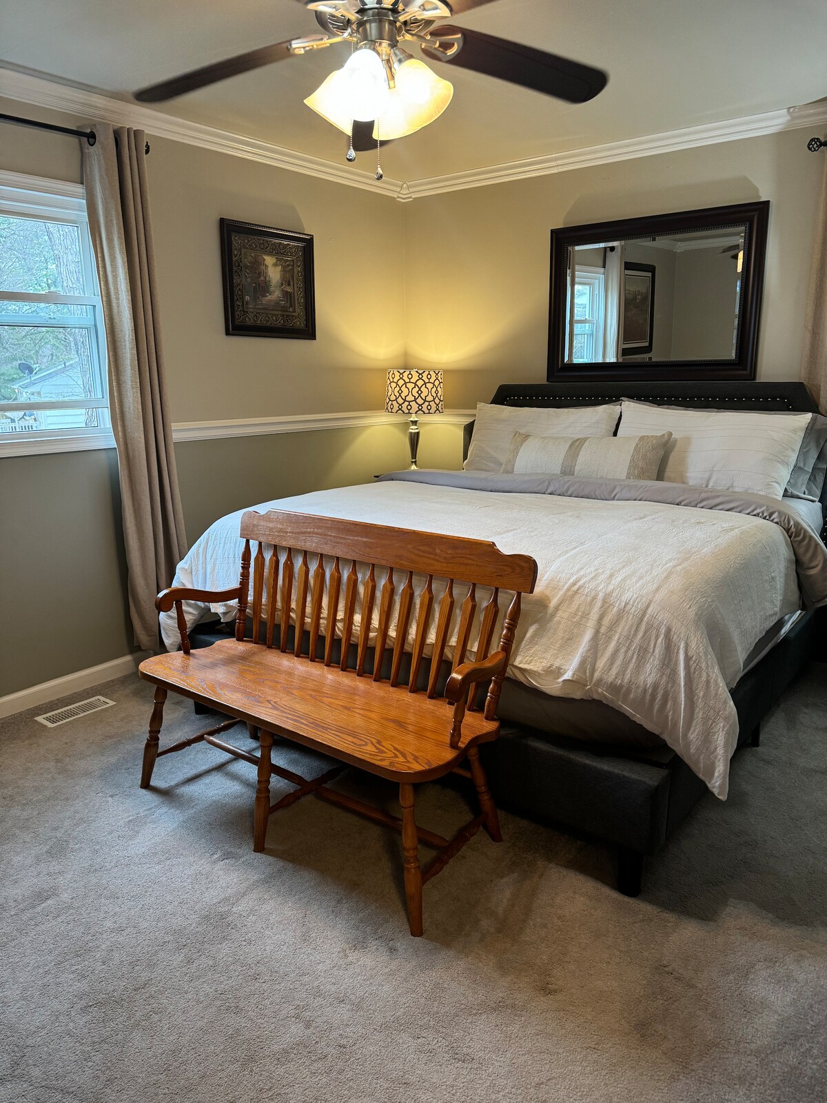 The bedroom features a queen-sized bed dressed in neutral linens, complemented by decorative pillows. A wooden bench is positioned at the foot of the bed, and a ceiling fan is visible above. Soft light is provided by a lamp beside the bed, enhancing the serene atmosphere.