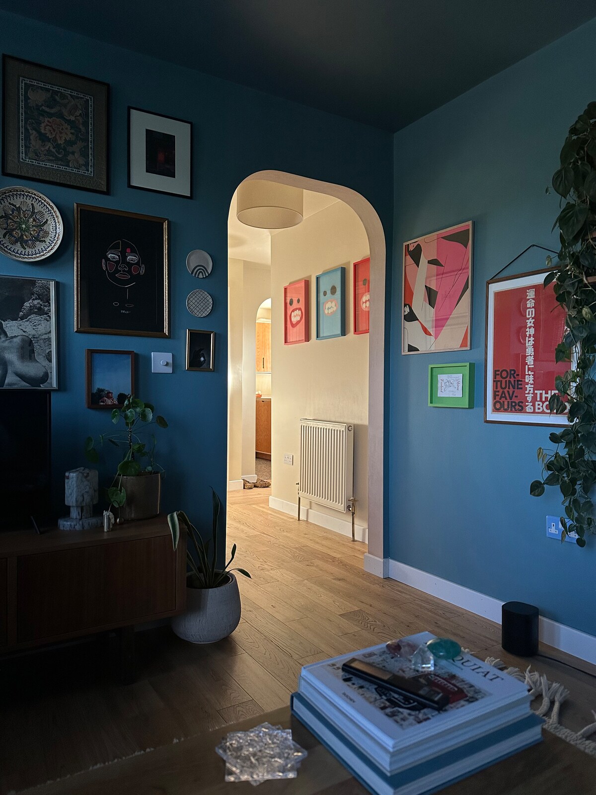 An inviting interior showcases a cozy lounge area with a large television and wooden console table. A doorway leads to a brightly lit hallway adorned with framed artwork. Soft lighting from a pendant fixture creates a welcoming atmosphere, complemented by a leaning plant by the wall.