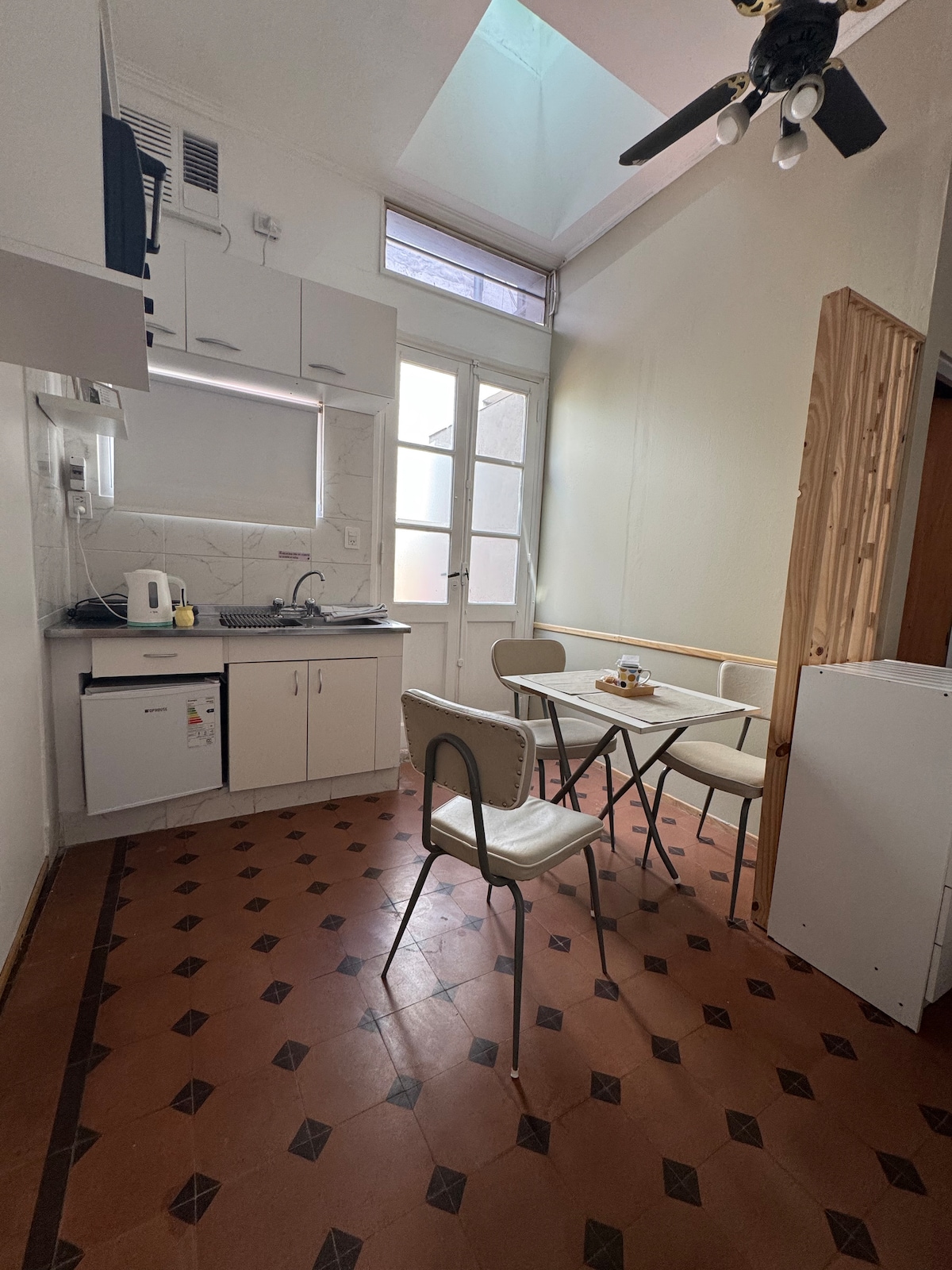 A compact kitchen area features white cabinetry and a tiled floor with a geometric pattern. A small dining table is set with tableware, surrounded by four light-colored chairs. Natural light enters through nearby windows, enhancing the cozy atmosphere.