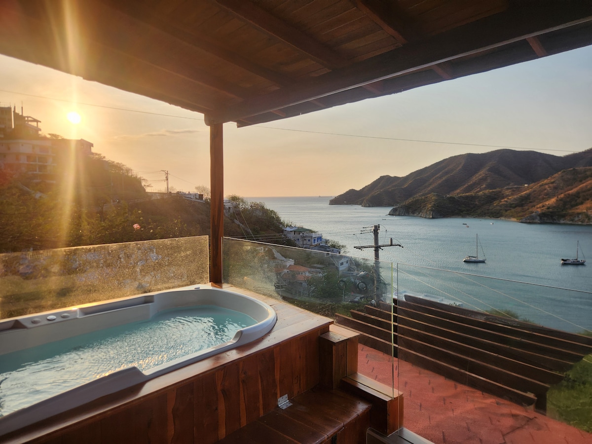 A private terrace features a jacuzzi overlooking the bay, with a panoramic view of the water and hills. Soft sunlight filters through, creating a tranquil ambiance. Wooden accents frame the space, enhancing the connection with nature.
